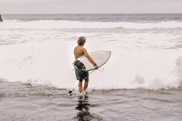 Back view of young fit surfer man with surfboard runs into ocean or sea with big waves for surfing. Concept of extreme sport, fitness, freedom, happiness, new modern life. Sports travel destination - Foto, Imagem