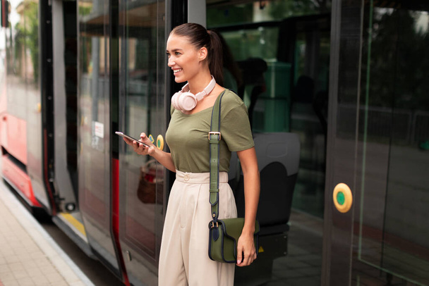 電車を降り、外に出て外を見ながら電話を使って幸せな乗客の女性。市内の公共交通機関で働くために通勤しながら、若い女性のテキストとウェブサーフィン - 写真・画像