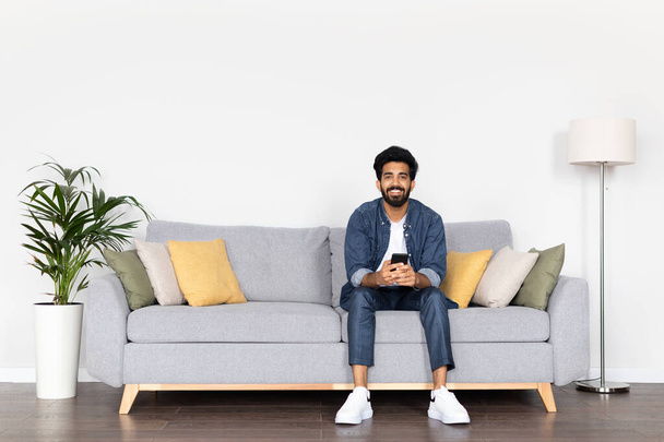 自宅のソファでリラックスしながら、スマートフォンを使用して幸せなアラブ人の男。新しいアプリを閲覧したり、オンラインで仕事をしたり、インターネットで買い物をしたり、リビングルームに座って無料のスペース - 写真・画像