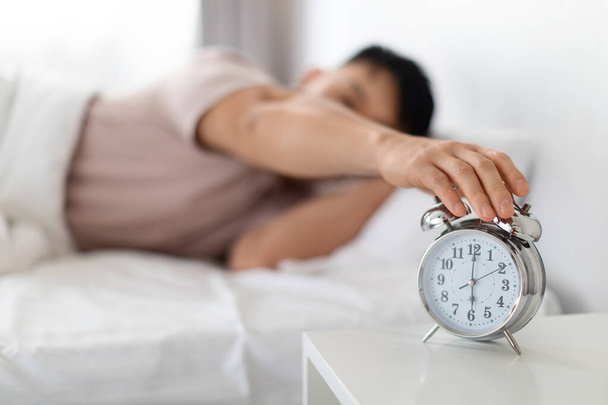 Wach auf, guten Morgen. Asiatischer Mann im Schlafanzug, der klingelnde Wecker ausschaltet, steht auf dem Nachttisch, wacht morgens auf, selektiver Fokus auf die Uhr, verschwommener Hintergrund - Foto, Bild