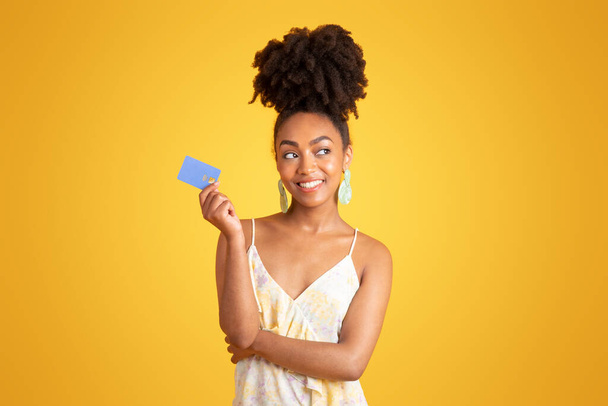 Cheerful pensive young black woman show credit card, dreaming about buying, isolated on yellow background, studio. Ad and offer, lifestyle, sale for shopaholic, pay for purchases - Photo, image