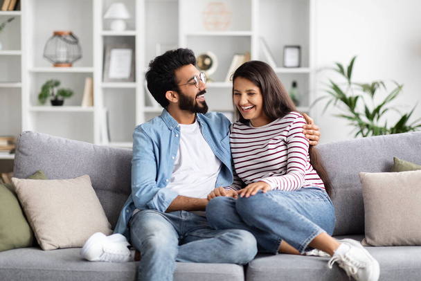 Szczęśliwy pozytywny piękny kochający młody indyjski para mężczyzna i kobieta spędzają czas razem w domu, siedzi na kanapie w przytulnym salonie, obejmując i śmiejąc się, kochankowie prowadzą rozmowę - Zdjęcie, obraz