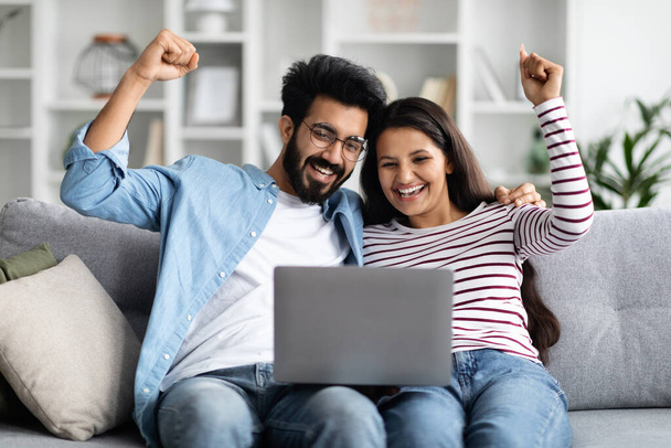 Emotional happy young beautiful indian couple wearing casual comfy outfits sittng on couch, using computer laptop at home, celebrating success. Gambling online, trading on stocks and markets - Foto, Bild