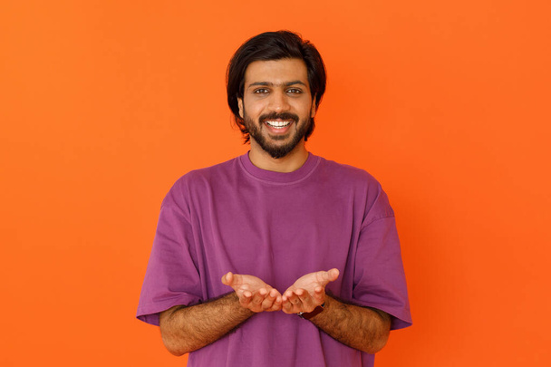 Happy smiling young indian man wearing casual purple t-shirt holding open hands next to his chest, hindu guy showing something invisible in his palms, isolated on orange studio background - Photo, Image