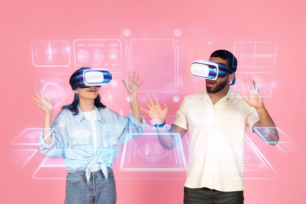 Excited joyful young couple experiencing virtual reality together, using wireless VR goggles, touching video game or simulator interface hologram, pink background, collage, double exposure - Photo, Image
