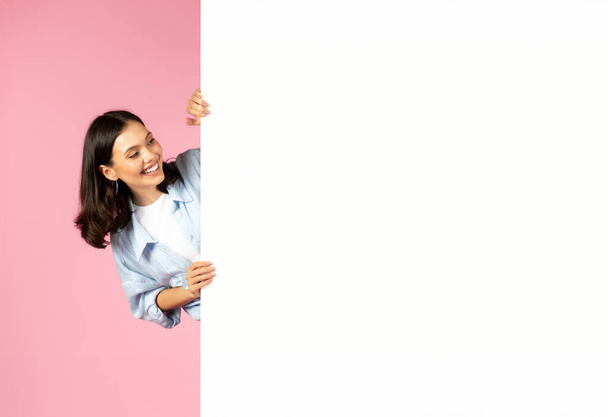 Opgewonden nieuwsgierig blij jong mooi brunette dame dragen casual outfit kijken naar enorme witte lege plakkaat mockup lege ruimte voor reclame, roze achtergrond. Zomer deal - Foto, afbeelding