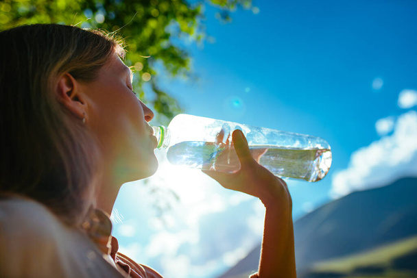 Young beautiful woman drinking pure water from a bottle in the mountains, focus on bottle - Photo, image