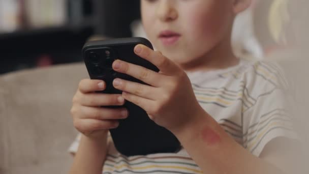 Happy small kid playing on mobile phone, watching video, sitting in armchair in living room interior. Addiction. App for entertainment and fun. - Footage, Video