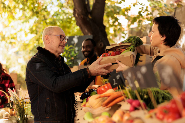 Senior άνθρωπος αγοράζει διάφορα φυσικά αγροτικά προϊόντα στο περίπτερο των αγροτών, πωλητής δίνοντας κουτί με βιολογικά φρέσκα προϊόντα. Γέρος που καλλιεργεί πολύχρωμα οικολογικά φρούτα και λαχανικά. - Φωτογραφία, εικόνα
