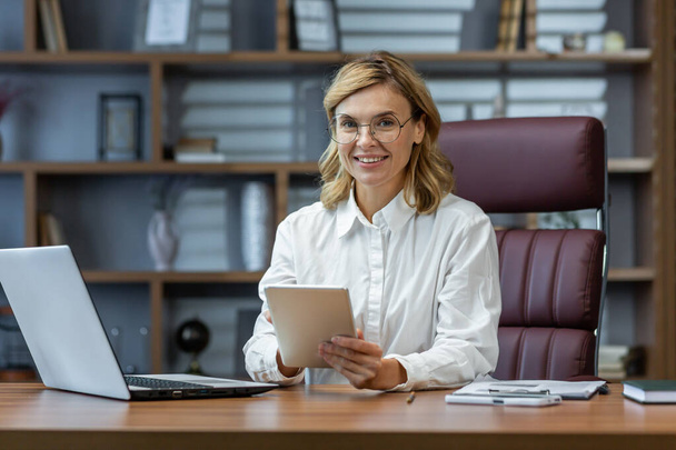 Ώριμη έμπειρη γυναίκα που εργάζεται μέσα στο γραφείο στο χώρο εργασίας, πορτρέτο της επιχειρηματία κρατώντας tablet υπολογιστή, αφεντικό χαμογελά και κοιτάζοντας κάμερα. - Φωτογραφία, εικόνα