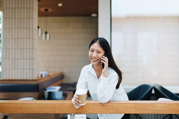 Una giovane asiatica lavoratrice autonoma che mostra un volto sorridente e felicità mentre usa smartphone mobile parlando d'affari e bevendo caffè in un bar. lavoratore autonomo, freelance, relax - Foto, immagini