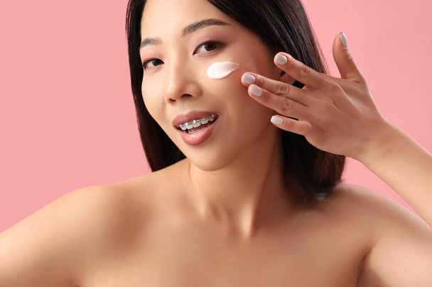 Красивая азиатка со сливками на лице на розовом фоне - Фото, изображение