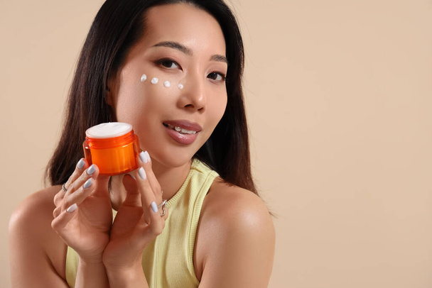 Schöne asiatische Frau mit Gesichtscreme auf beigem Hintergrund - Foto, Bild