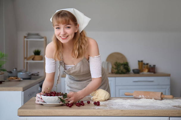 Beautiful young woman with bowl of cherries, rolling pin and dough on table in kitchen - Фото, изображение