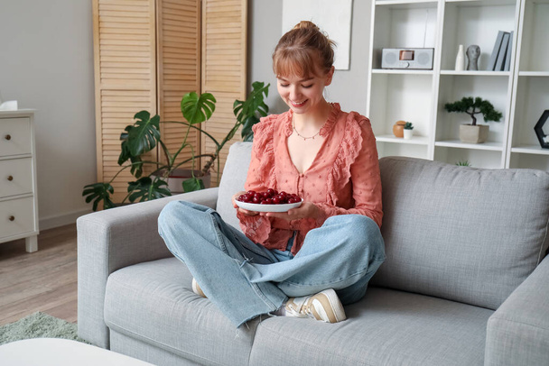 Hermosa joven feliz con un tazón lleno de cerezas maduras sentadas en el sofá en la sala de estar - Foto, imagen