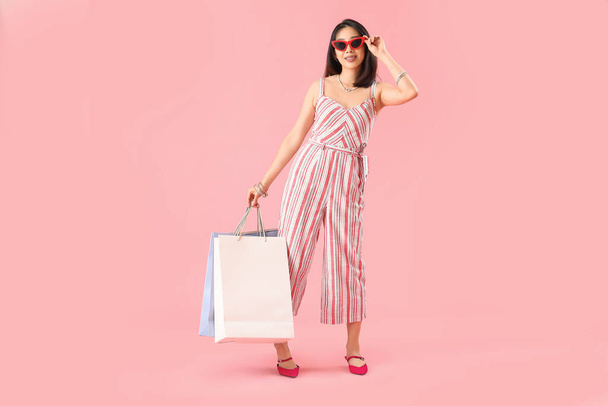 Glücklich schöne junge Asiatin in Sonnenbrille mit Einkaufstaschen auf rosa Hintergrund - Foto, Bild