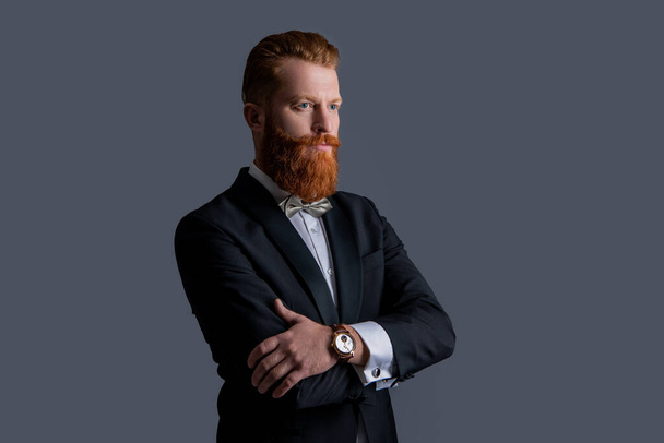man studio shot in tuxedo and wristwatch. man wearing elegant wristwatch and tuxedo. tuxedo fashion. handsome bearded man in tuxedo with wristwatch isolated on grey background. - Foto, afbeelding