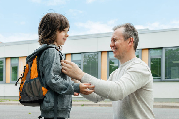 A father helps his son to put on a backpack. The child is happy to go back to school or kindergarten. Parents take care of children - Foto, imagen