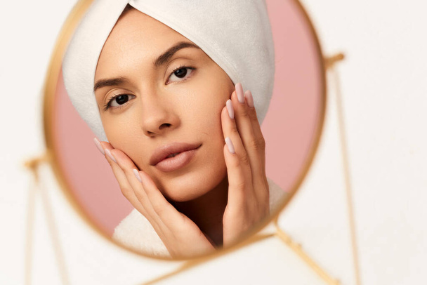 View in round mirror of young female in white bathrobe and towel on head touching face while applying cosmetic product during skincare routine - Photo, Image