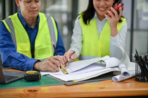 Dos ingenieros de construcción asiáticos profesionales que usan chalecos de seguridad están revisando un plan, discutiendo el trabajo y trabajando juntos en la oficina. - Foto, Imagen