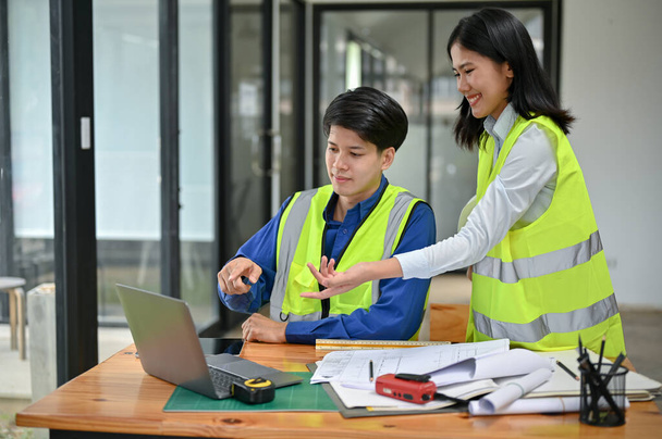 Un ingeniero masculino asiático inteligente y profesional en un chaleco de seguridad está mirando una pantalla de computadora portátil y discutiendo un plano del edificio en el escritorio con su colega femenina en la oficina. - Foto, Imagen