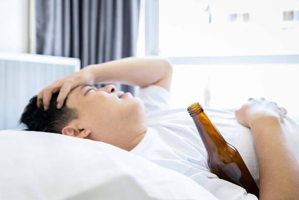 Kocovina muž trpí těžkou bolestí hlavy v dopoledních hodinách venku pití, Opilý muž středního věku objímající láhev piva, ležící na posteli, mající kocovinu, bolest v hlavě, pití přebytek alkoholu - Fotografie, Obrázek