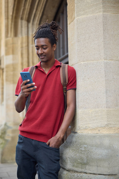 Male Student Checking Messages Or Social Media On Mobile Phone Outside University Building In Oxford UK - Zdjęcie, obraz