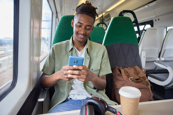 Young Man Commuting To Work On Train Sitting  On Train Looking At Mobile Phone For Travel Information Or Social Media - 写真・画像