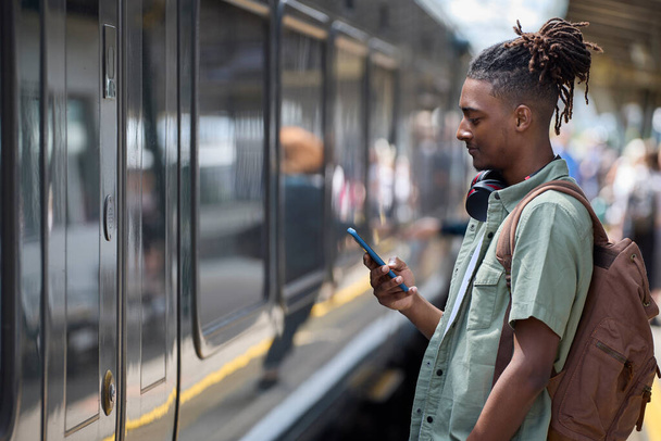 Young Man Commuting To Work On Train Standing  On Platform Looking At Mobile Phone As Train Arrives - Foto, imagen