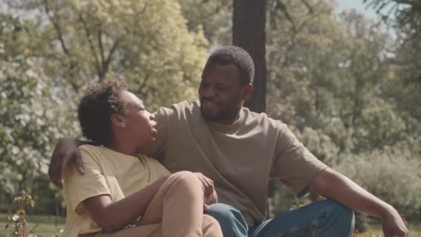 Średnie spowolnienie zrelaksowanego Afroamerykanina ze swoim 12-letnim synem siedzącym na zielonej trawie w parku, cieszącym się naturą i mówiącym - Materiał filmowy, wideo