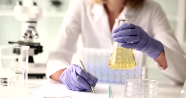 Female lab worker holds flask with examined urine of patient writing down results of research. Lab worker at sterile clinic workplace writes data into medical blank - Footage, Video