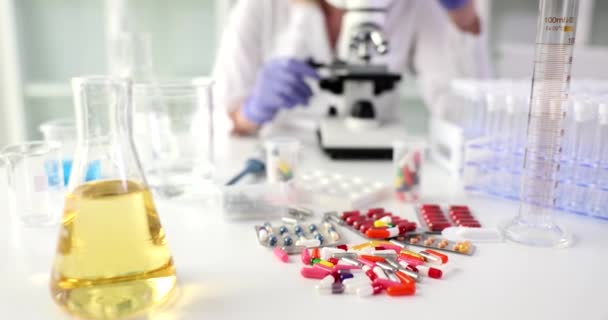 Frasco de orina en mesa de laboratorio con pila de pastillas contra científicos que trabajan con microscopio. Tratamiento de la enfermedad del sistema urinario con medicamentos. Investigación de la utilidad de las píldoras - Imágenes, Vídeo