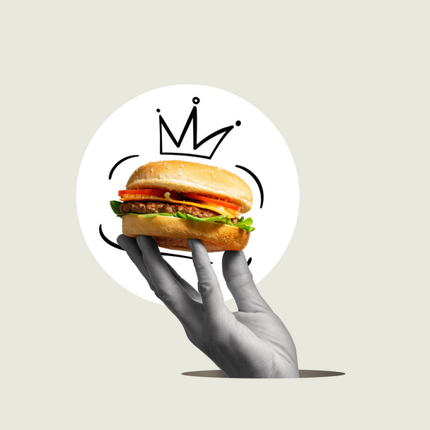Een hand houdt een hamburger vast met een handgetekende kroon. Kunstcollage. - Foto, afbeelding