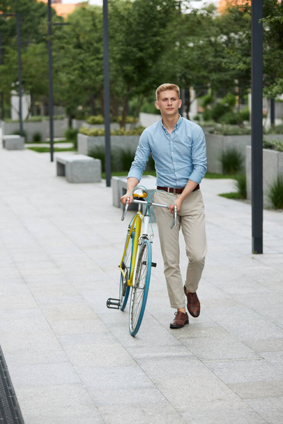Full-length image of young handsome man, employee in smart casual clothes walking along the street with bicycle on early summer morning. Concept of business, active lifestyle, fashion, youth, ecology - Foto, afbeelding