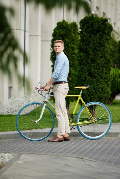 Handsome, serious young man, employee standing in stylish formal clothes on street near office building with bicycle. Going to work. Concept of business, active lifestyle, fashion, youth, ecology - Foto, Bild