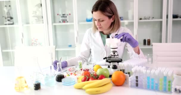 女性科学者は顕微鏡を使って果物の物質の構造を調べ、結果を空白に書き込みます。果物の装置と錠剤の山とテーブルのラボの労働者 - 映像、動画