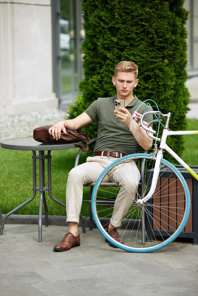 Stylish, handsome, serious businessman sitting on bench and reading news, mails on mobile phone. Going to work on bike. Concept of business, active lifestyle, fashion, youth, ecology - Valokuva, kuva