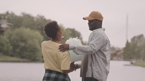 Vue arrière slow moyen de l'homme afro-américain et son fils de 12 ans avec des barbe à papa à la main debout au bord de la rivière et parler, profiter de la nature et du temps ensemble - Séquence, vidéo