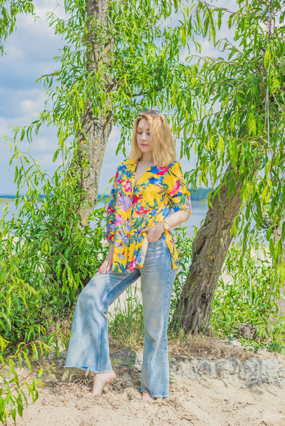 Styl wakacyjny, kobieca garderoba. Ilustracja młodej europejskiej opalonej kobiety z blond włosami w niebieskich dżinsach i kolorową bawełnianą koszulą na plaży - Zdjęcie, obraz