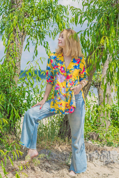 Estilo de vacaciones, armario de mujer. Ilustración de una joven bronceada europea con cabello rubio en jeans azules y camisa de algodón colorido en la playa - Foto, Imagen