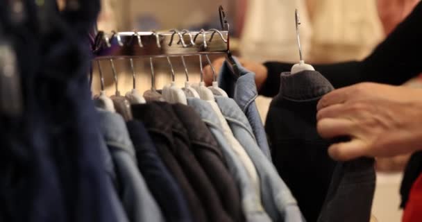 Człowiek wybiera pomiędzy czarnymi i niebieskimi kurtkami jeansowymi stojąc przed stoiskiem pełnym płaszczy w sklepie. Człowiek dokonuje wyboru na kurtkę kupić - Materiał filmowy, wideo