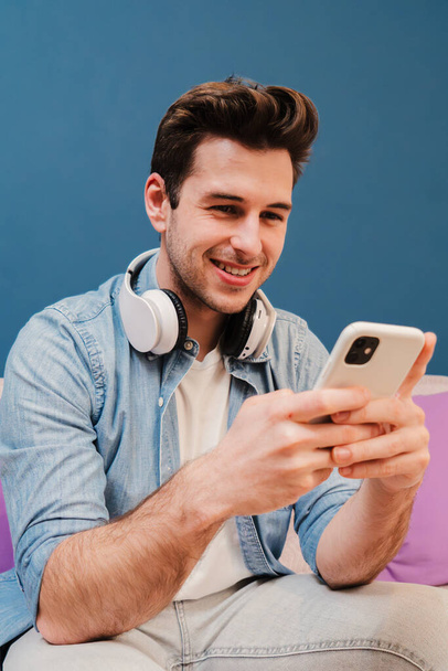 Κάθετη πορτρέτο ενός εφήβου που περνάει καλά στο διαδίκτυο με ένα κινητό. Νεαρός χρησιμοποιεί ένα smartphone κάθεται στον καναπέ στο σπίτι σαλόνι. Guy χαμογελώντας και μοιράζονται στην εφαρμογή κοινωνικών μέσων μαζικής ενημέρωσης - Φωτογραφία, εικόνα