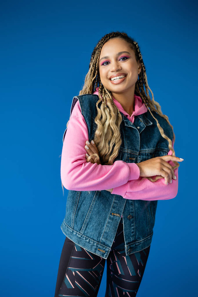 joyful african american woman in pink hoodie and denim vest on blue background, bold makeup - Photo, Image