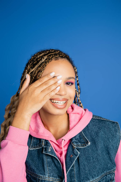 mujer feliz con rastas cubriendo el ojo y mirando a la cámara en el fondo azul, maquillaje en negrita - Foto, imagen