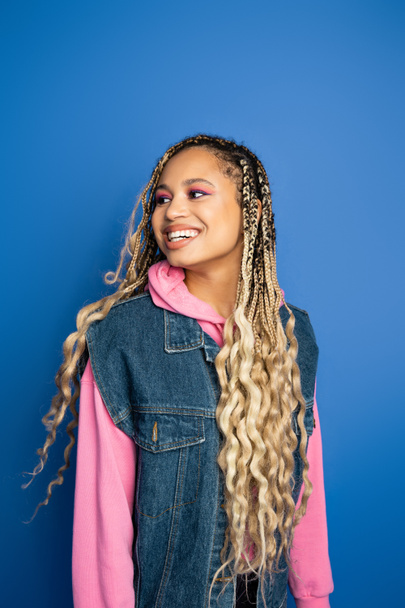 joyful african american woman with dreadlocks smiling on blue background, denim vest and hoodie - Photo, Image