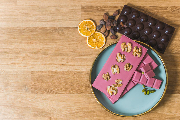 Serving suggestion concept. Brs of ruby aand dark chocolate arrenged on blue plate and wooden table decorated with walnuts slices of orange and cocoa seeds. Copy space. High quality photo - Photo, Image