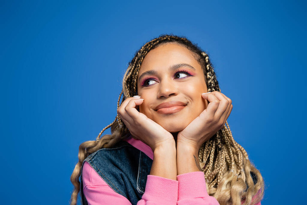 portrait of happy and dreamy african american woman with bold makeup looking away on blue background - Photo, image