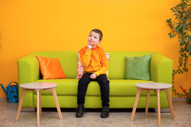 Cute pensive child boy in a bright orange sweater sits on a green sofa - Photo, Image
