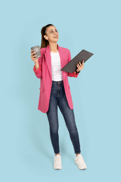 Full length business woman, cheerful young brunette full length business woman. Holding clipboard papers and coffee wearing wireless earphones. Smiling positive girl standing over blue background. - Photo, Image
