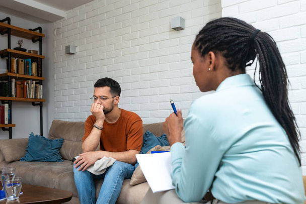 Young depressed man crying talking to mental health counselor during a session in the office. Stressed male at psychotherapy after divorce having emotional damage issue. Relationship difficulties. - Foto, afbeelding